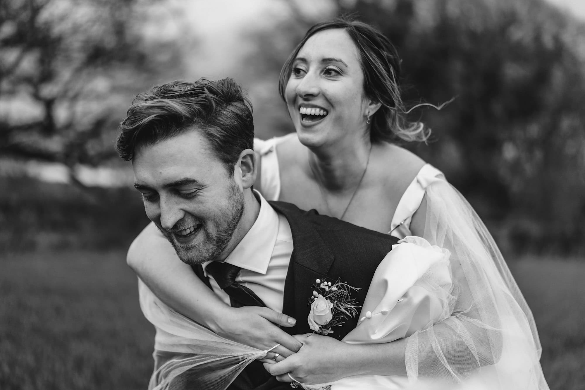 Bride riding piggy back on groom in the fields around Woodhall Manor